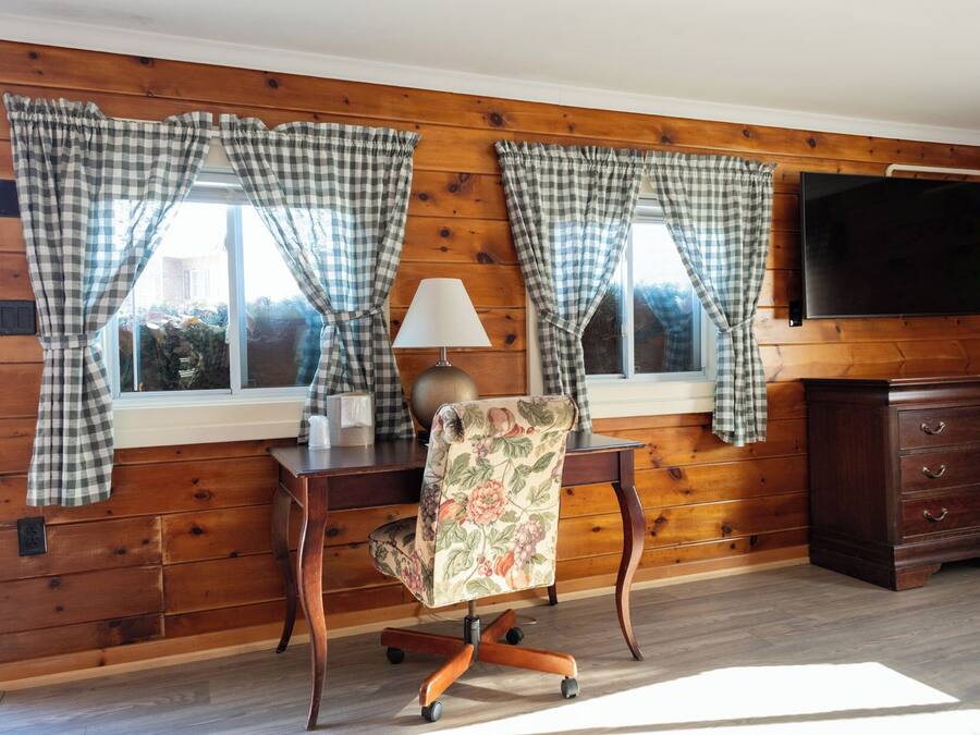 A room with wooden paneled walls, a desk with a lamp, a floral chair, and a TV mounted on the wall.