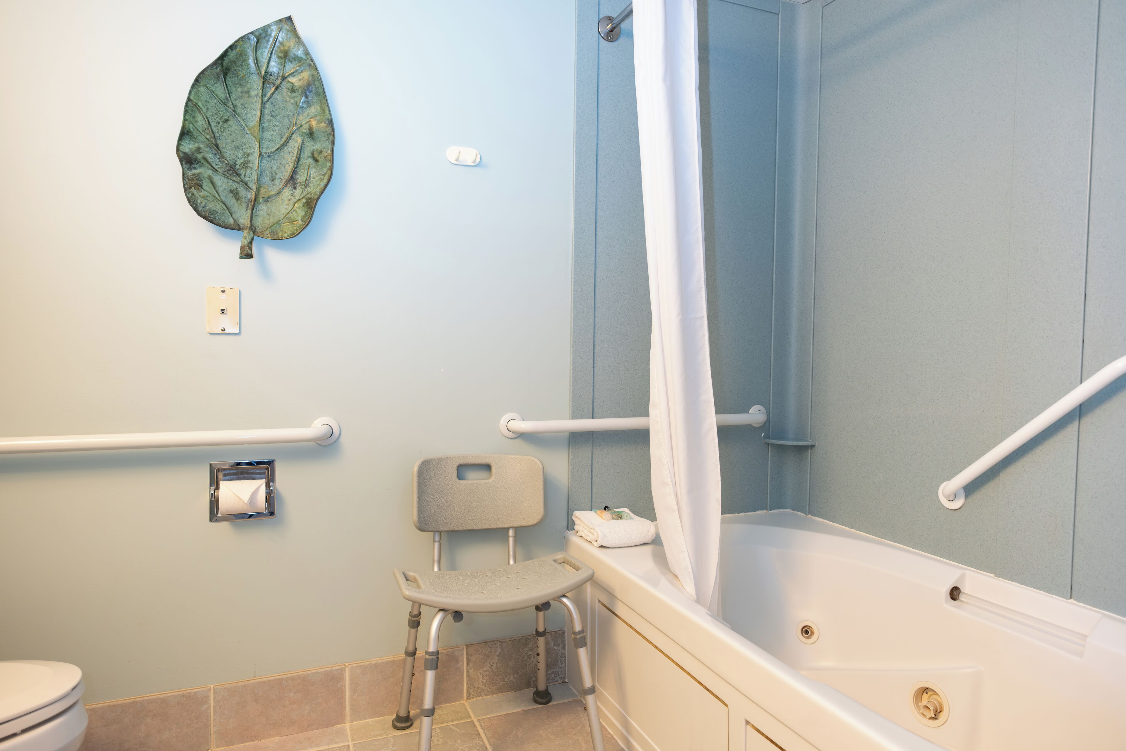 Bagno dotato di vasca, tenda da doccia, water, mancorrente e sedia accessibile in sedia a rotelle.
