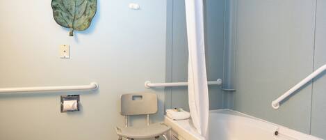 A bathroom with a bathtub, a shower curtain, a toilet, a handrail, and a wheelchair-accessible chair.
