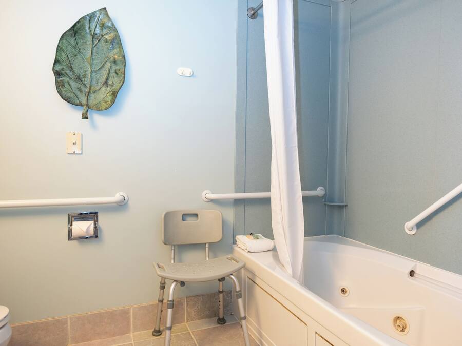 Bagno dotato di vasca, tenda da doccia, water, mancorrente e sedia accessibile in sedia a rotelle.