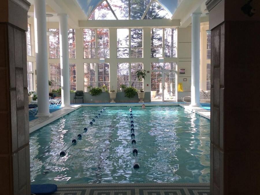An indoor swimming pool with clear glass walls, surrounded by columns and potted plants.