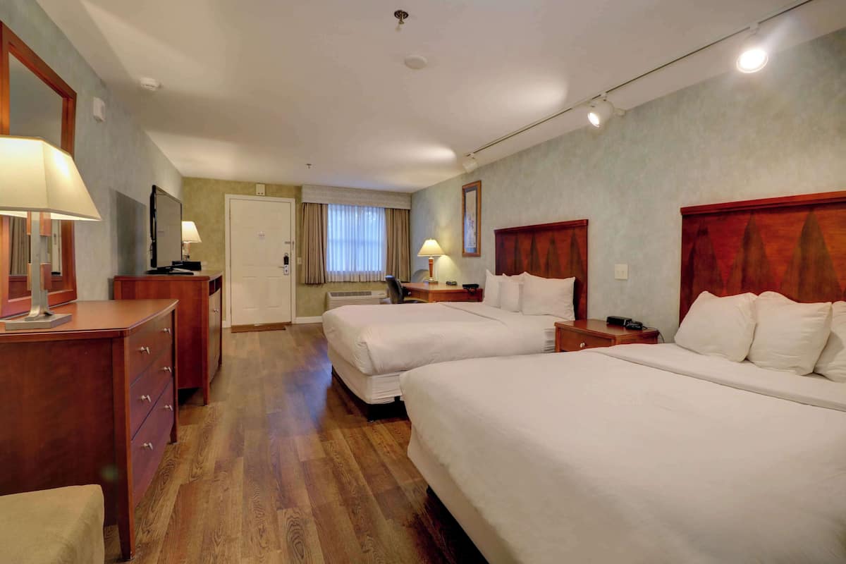 A hotel room with two beds, wooden headboards, a desk with a lamp, a dresser, and a large window with curtains.