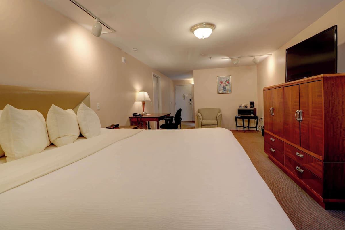 A hotel room with a large bed, a wooden wardrobe, a desk with a lamp, a chair, and a television.