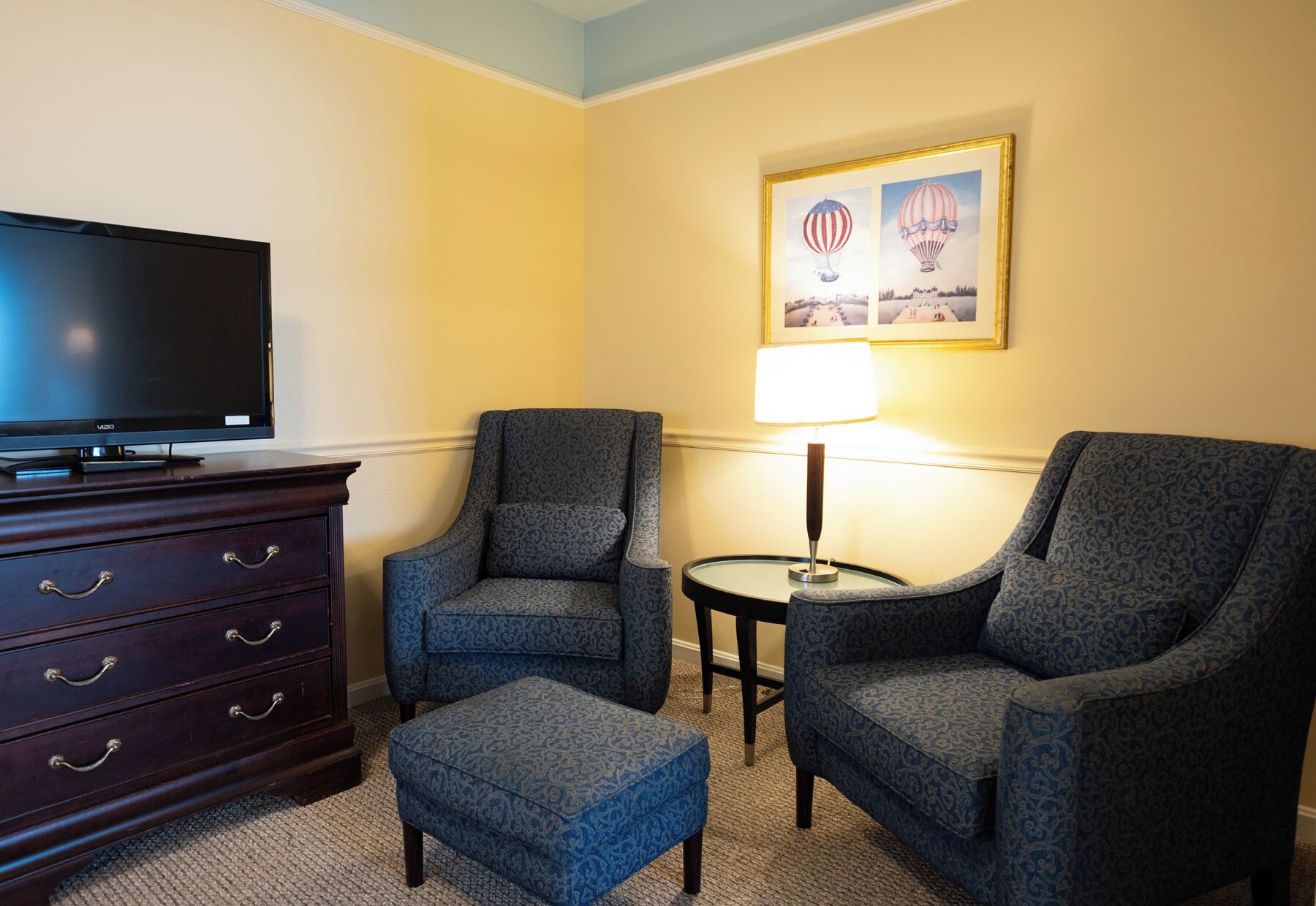 A hotel room with a flat-screen TV, two armchairs, a small ottoman, a lamp, and a framed picture of hot air balloons on the wall.
