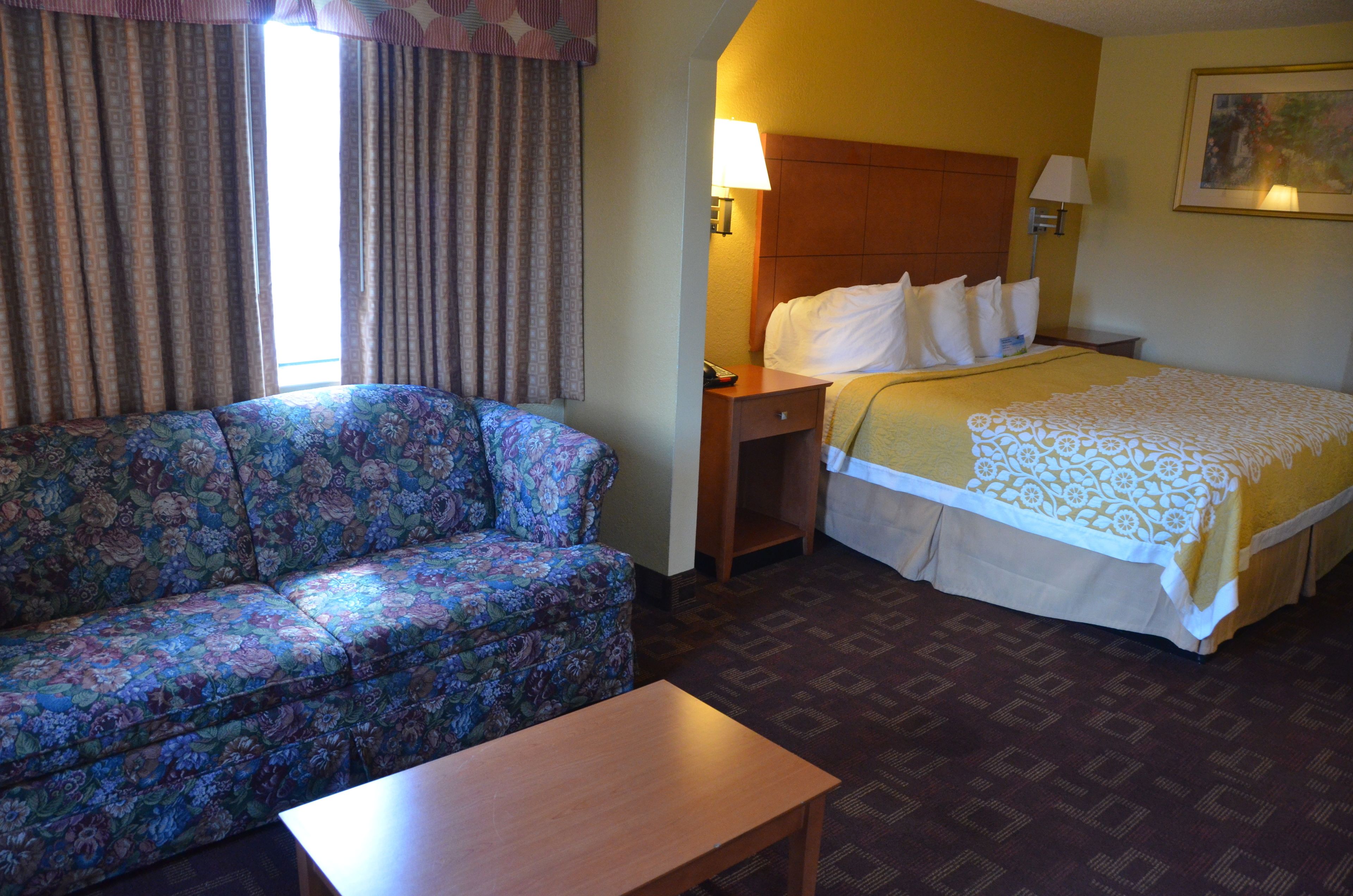 A hotel room with a floral sofa, a bed with a patterned bedspread, a nightstand with a lamp, and a wooden table.