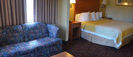 A hotel room with a floral sofa, a bed with a patterned bedspread, a nightstand with a lamp, and a wooden table.