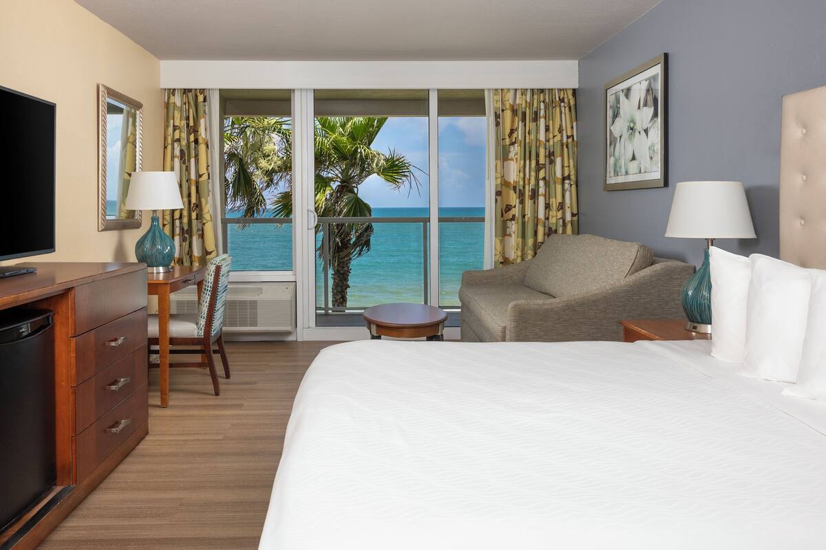 A hotel room with a large bed, a TV, a desk with a chair, a small table, and a view of the ocean through large windows.