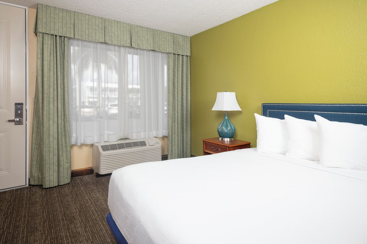 A hotel room with a large bed, a nightstand with a lamp, a window with sheer curtains, and a wall heater.