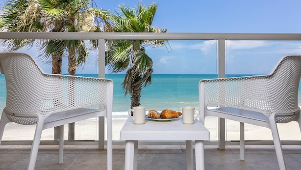 A balcony with two white wicker chairs, a small table, and a plate with croissants and two white mugs. The view includes a palm tree and the ocean. - Ocean Breeze Inn Vero Beach (Vero Beach)
