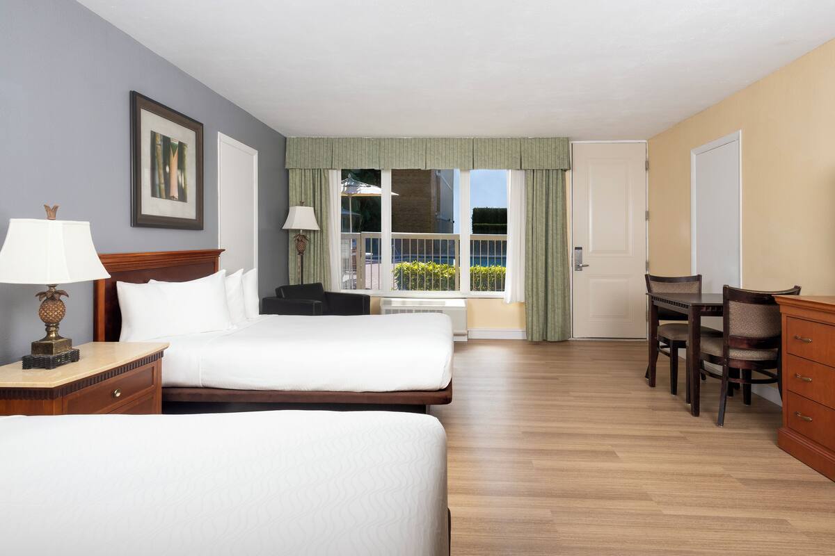 A hotel room with two beds, a desk, and a chair. There is a window with curtains, a lamp on the desk, and a picture on the wall.
