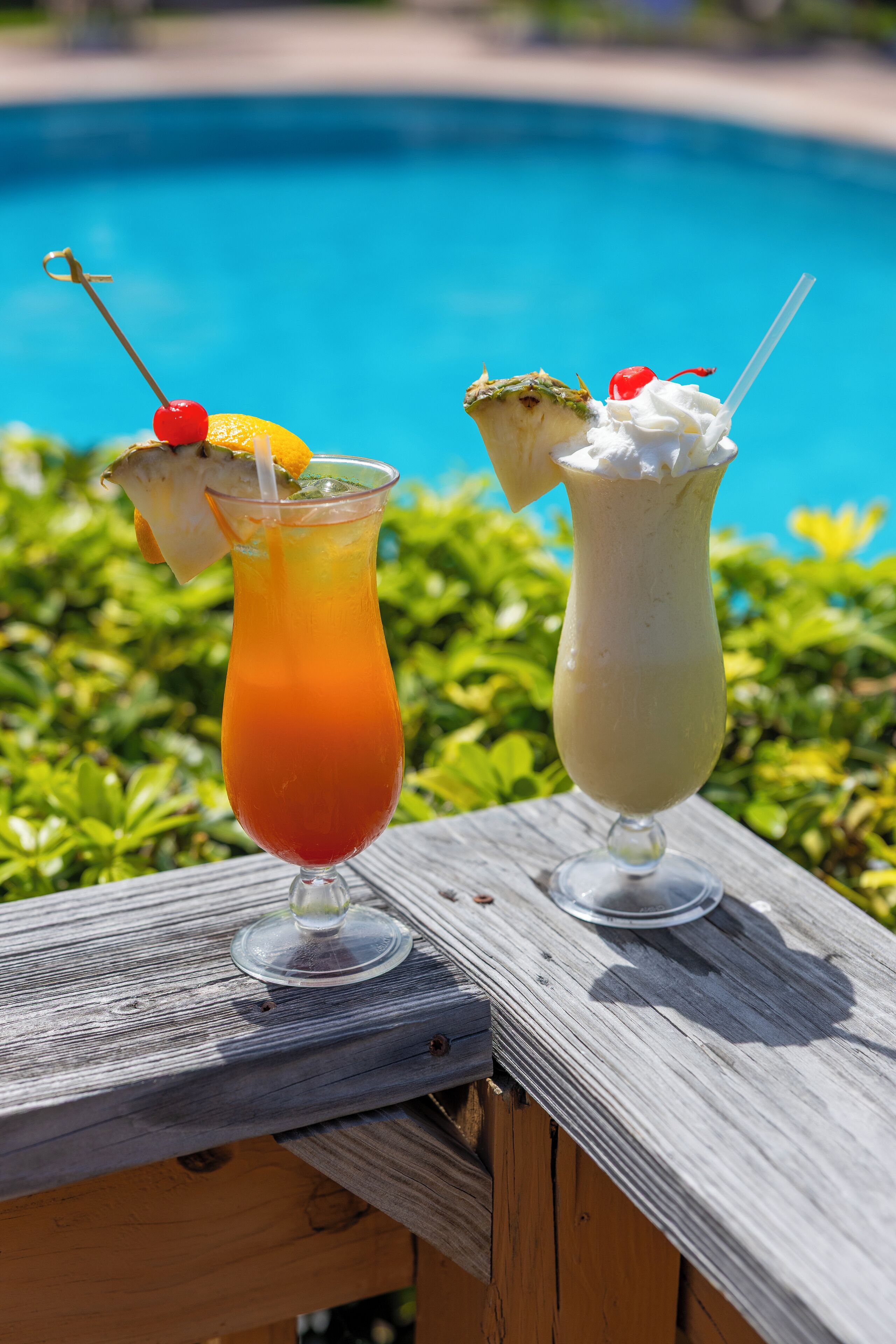 Two cocktails on a wooden table by a pool with a clear blue sky in the background.