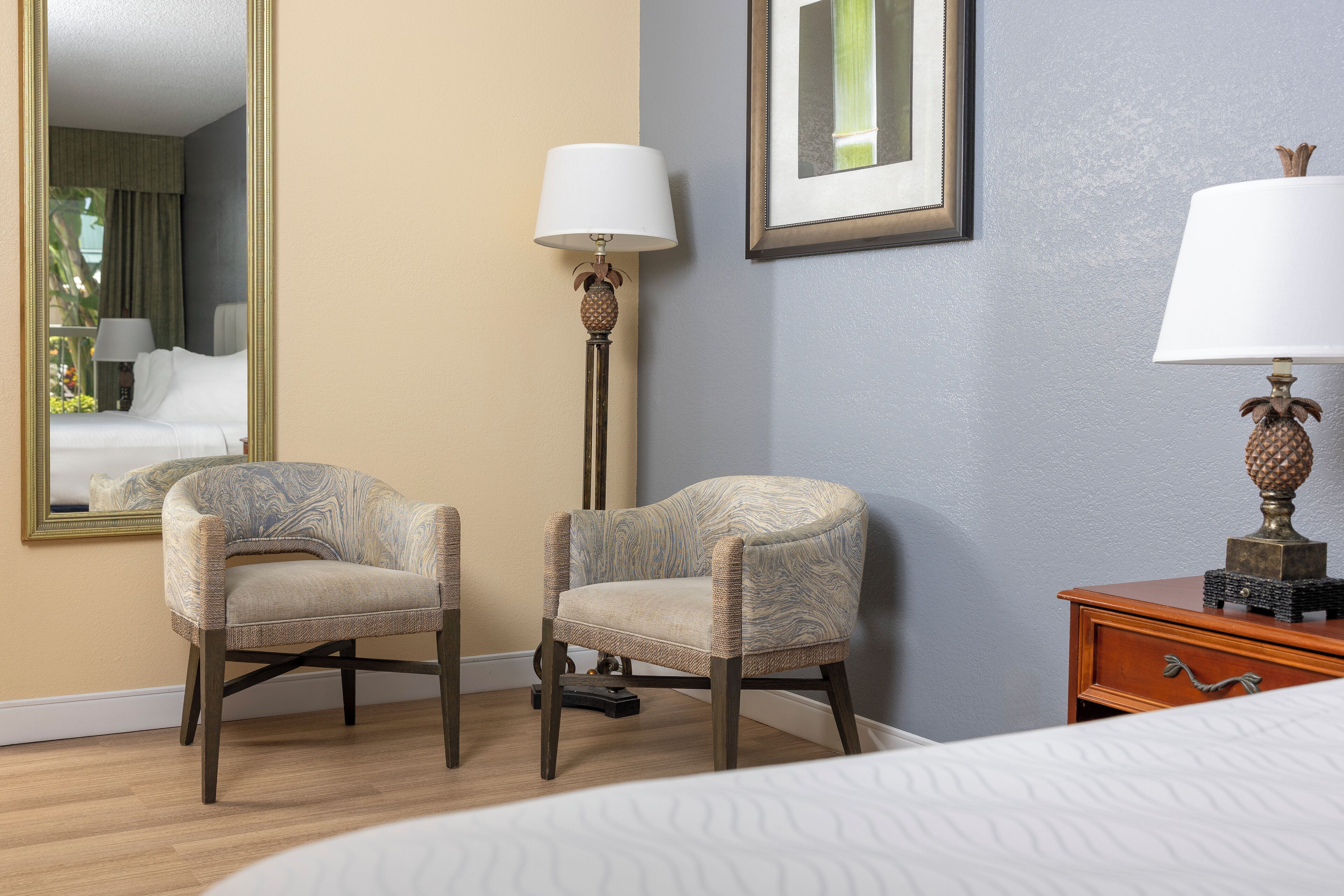 A hotel room with two armchairs, a floor lamp, a bedside table with a lamp, a bed, and a large mirror on the wall.