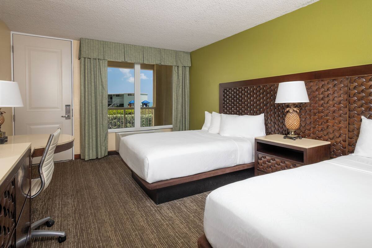 A hotel room with two beds, a desk with a chair, a window with curtains, and a view of the outside.