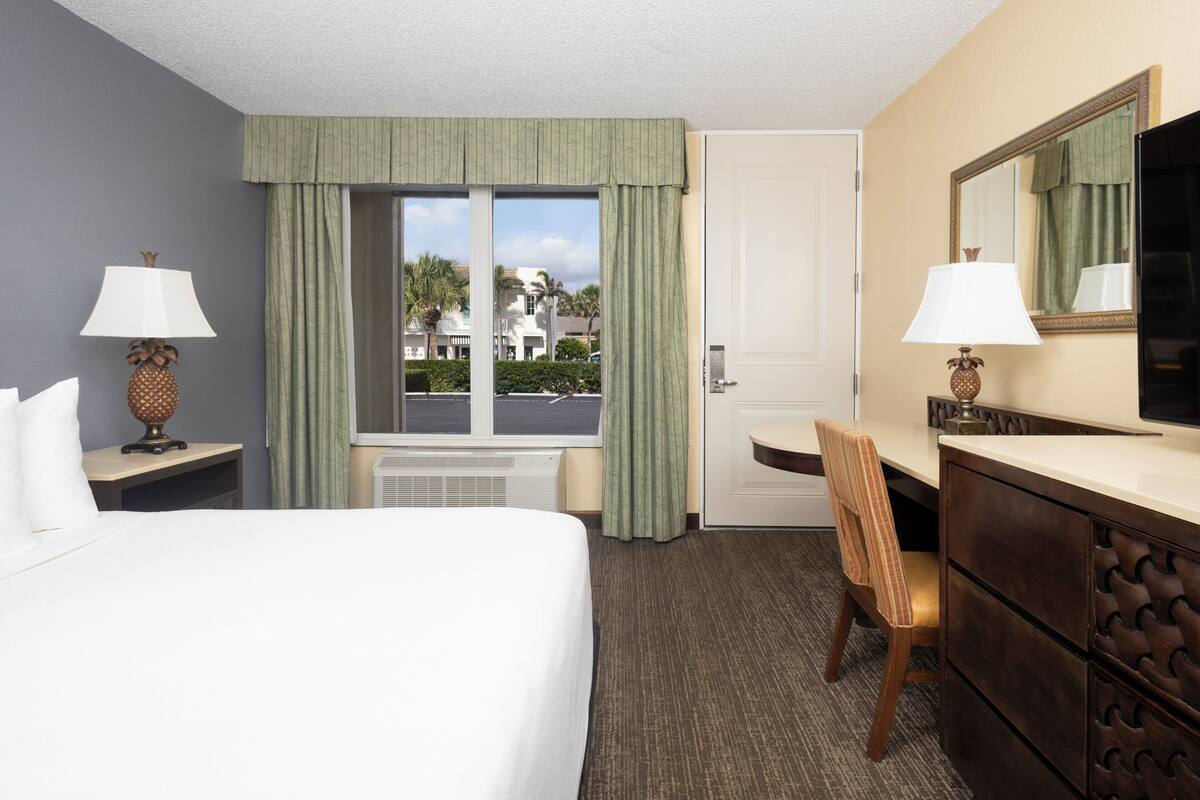 A hotel room with a bed, a desk, a chair, a dresser, a mirror, and a window with curtains.