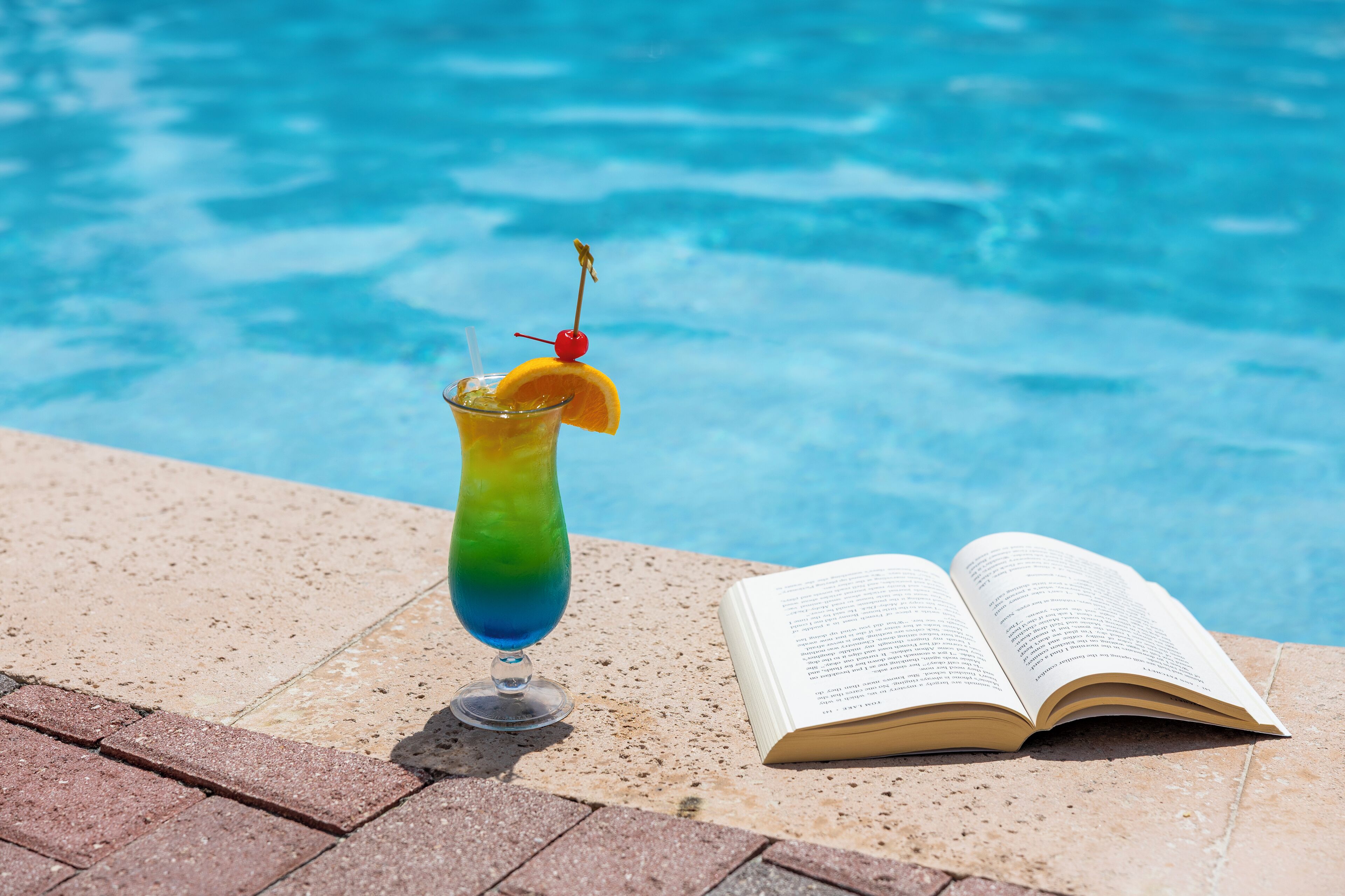 Um copo de vidro transparente com bebida degradê, um cereja espetada num palito e um livro aberto ao lado de uma piscina.