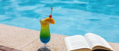 A clear glass with a gradient drink, a cherry on a stick, and an open book beside a pool.