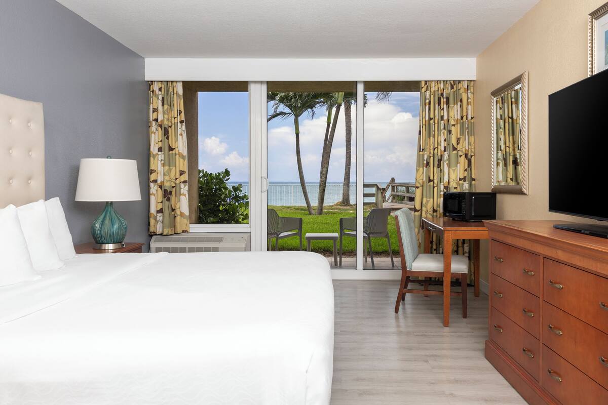 A hotel room with a large bed, a desk with a chair, a TV, and a view of the ocean through a sliding door.