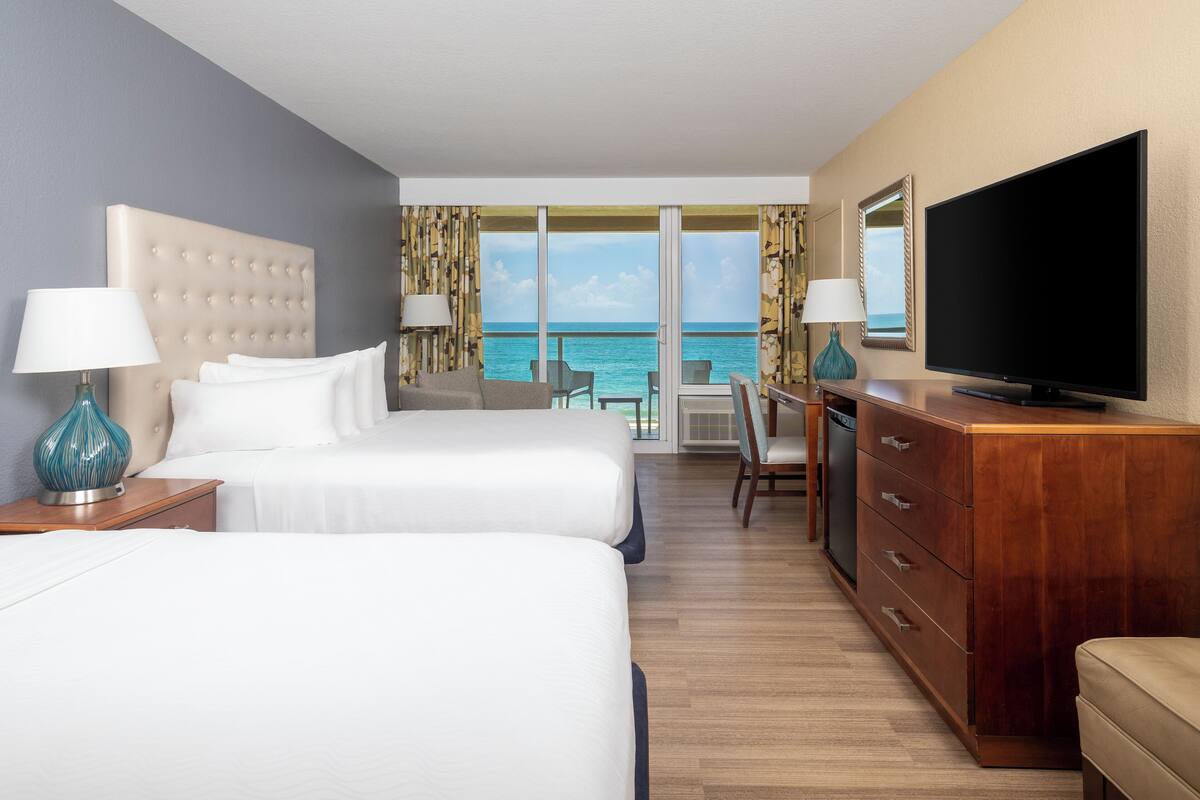 A hotel room with two beds, a TV, a desk, and a view of the ocean.