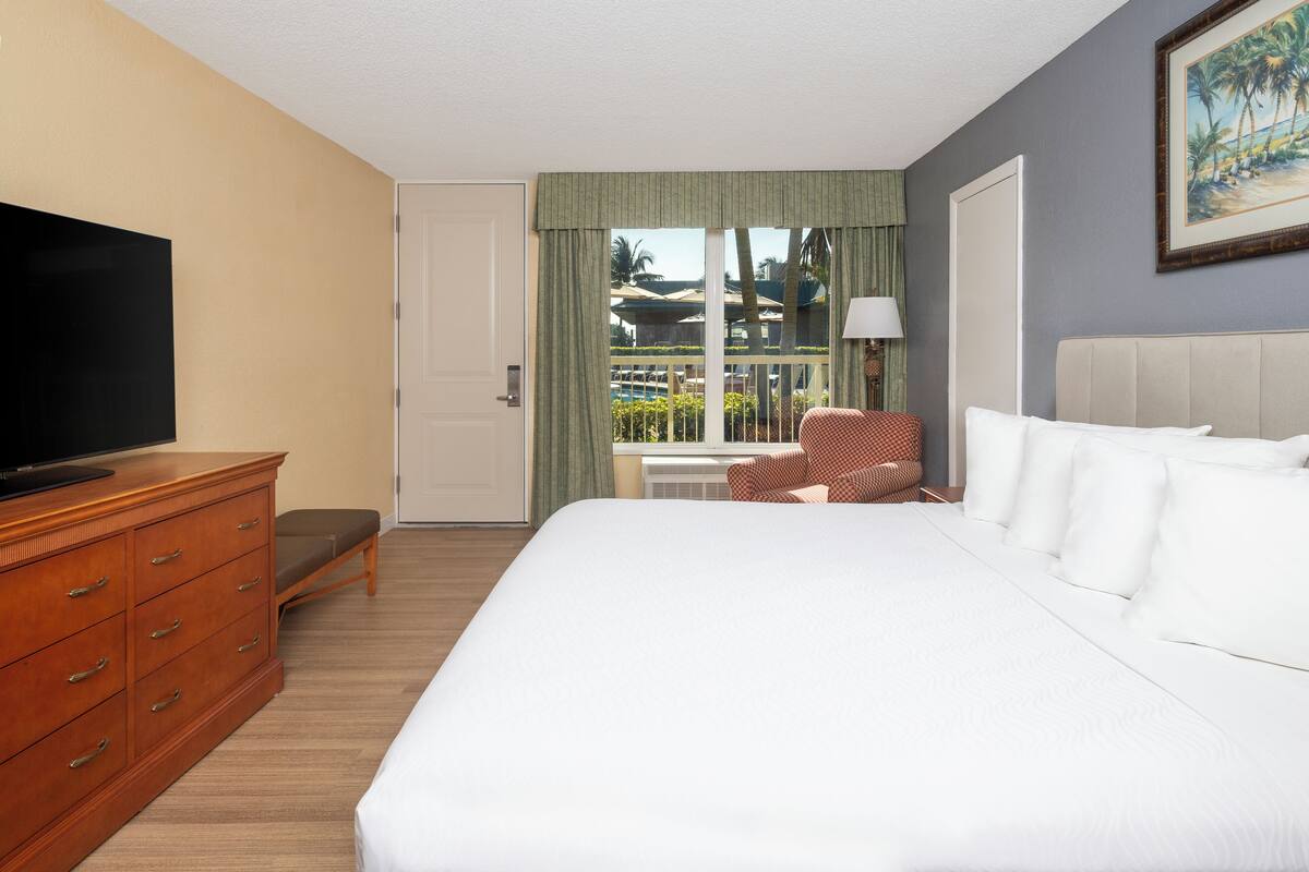 A hotel room with a large bed, a wooden dresser, a television, a chair, and a window with a view of palm trees and a pool.