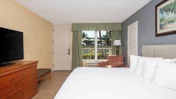 A hotel room with a large bed, a wooden dresser, a television, a chair, and a window with a view of palm trees and a pool.