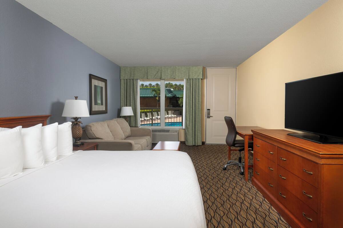 A hotel room with a bed, a desk, a television, a chair, and a view of a pool.