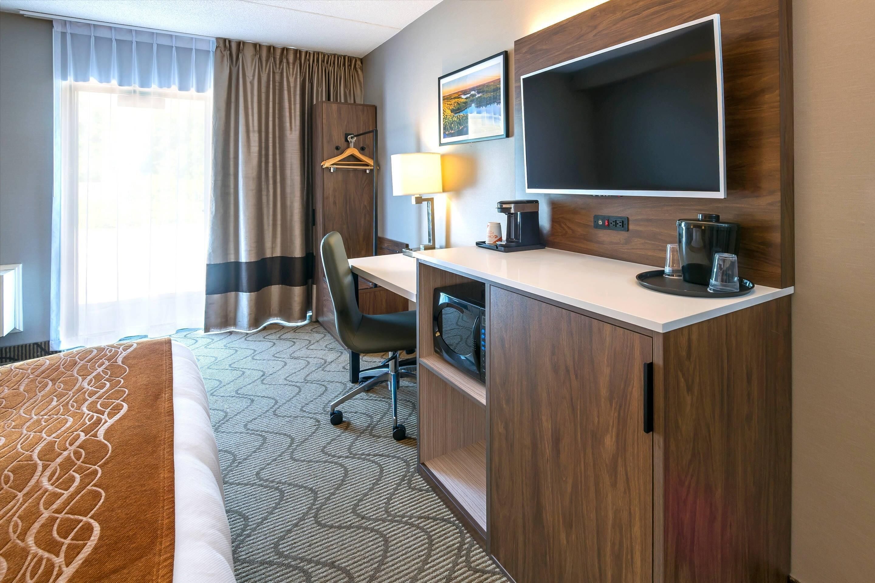 Een hotelkamer met een bed, bureau, stoel, lamp, koffiezetapparaat, televisie en een raam met gordijnen.