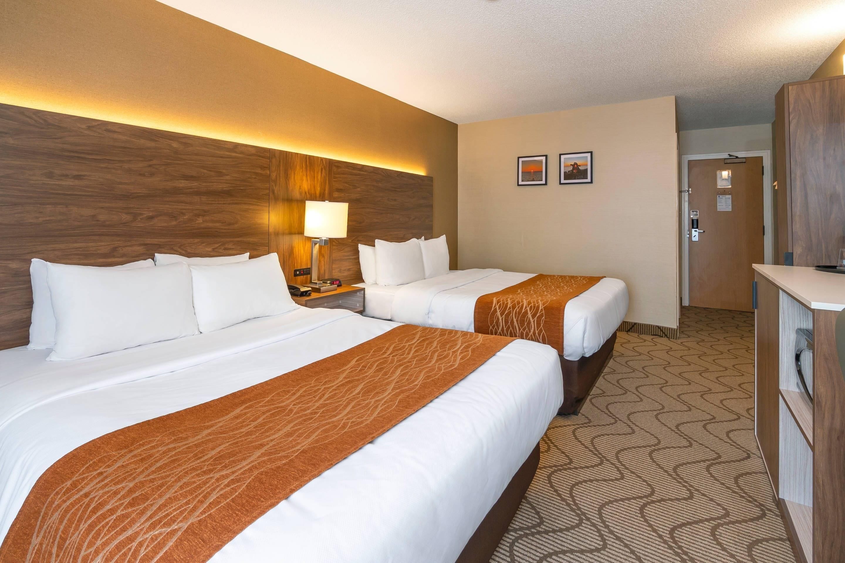 Habitación de hotel con dos camas, una mesita de noche con lámpara, cabecera de madera y puerta que da al baño.