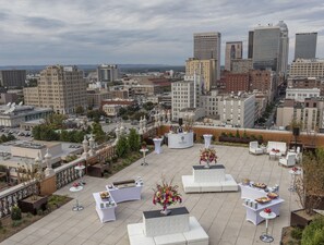 A rooftop terrace with white furniture and a buffet setup, overlooking a cityscape with various buildings.