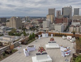 A rooftop terrace with white furniture and a buffet setup, overlooking a cityscape with various buildings.