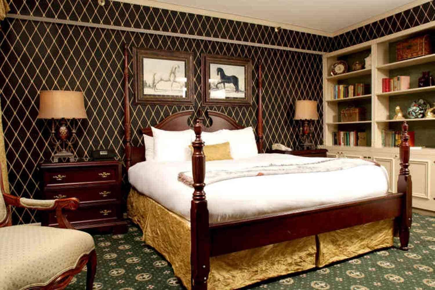 A four-poster bed with a green patterned carpet, a wooden nightstand with a lamp, a chair, and a bookshelf with books and decorative items.