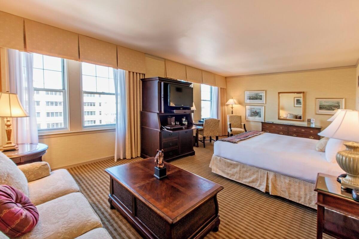 A hotel room with a large bed, a TV, a desk, and a chair. There are windows with curtains, a lamp, and a decorative item on the desk.