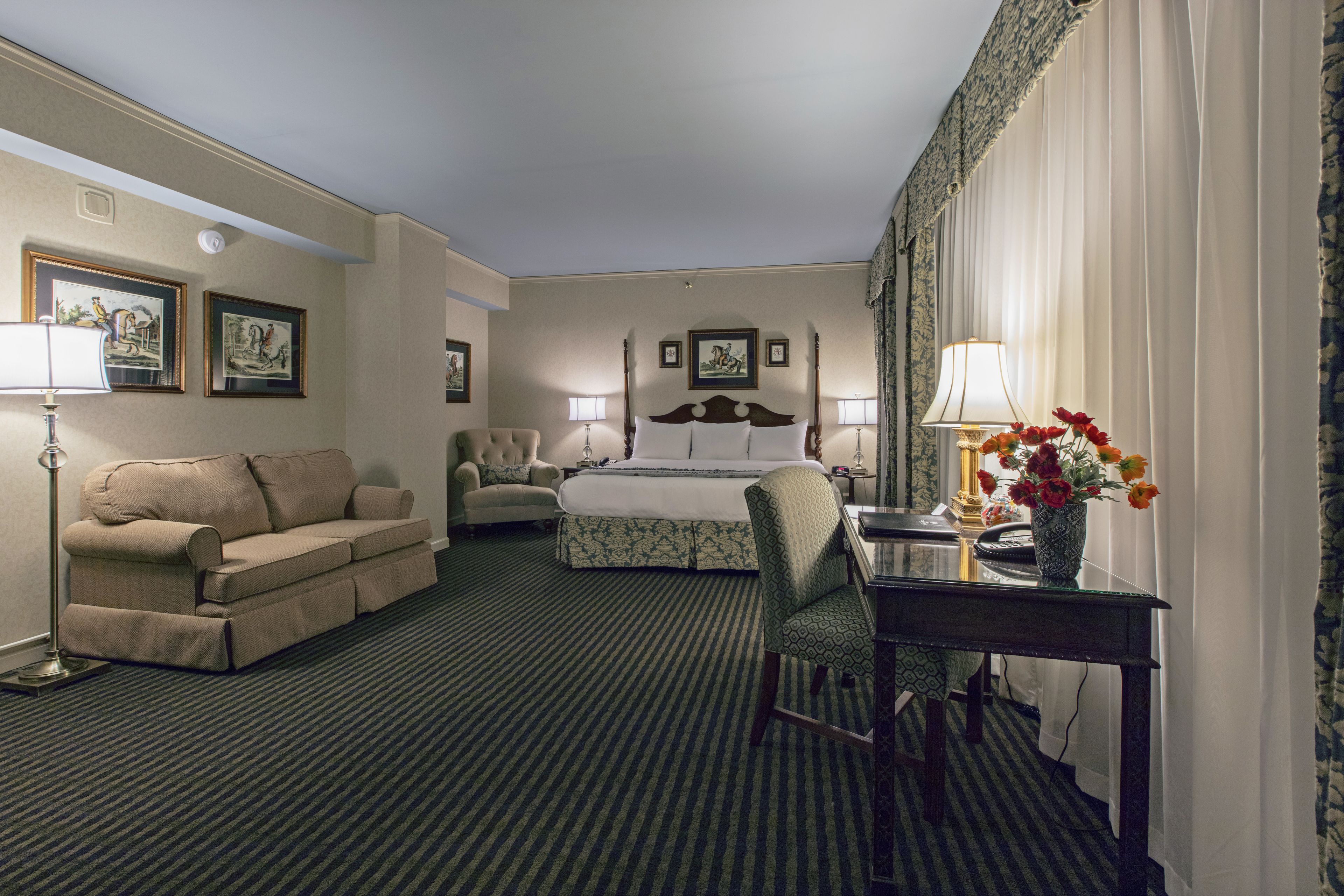 Habitación de hotel con cama, sofá, sillón, escritorio con lámpara y un florero con flores.