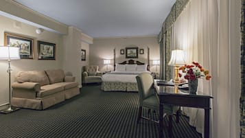 A hotel room with a bed, a sofa, a chair, a desk with a lamp, and a vase with flowers.