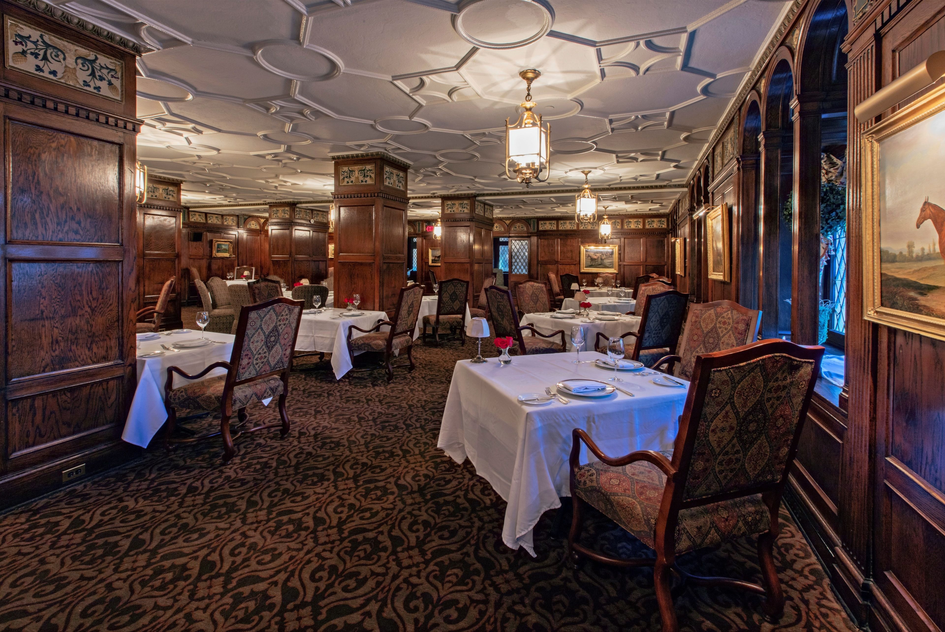 Un comedor formal con paneles de madera, techo ornamentado y candelabros. Las mesas están dispuestas con manteles blancos y sillas.