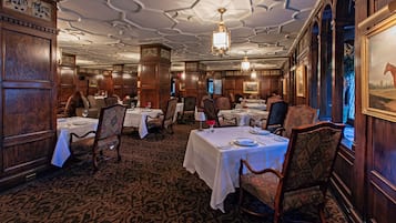 Un comedor formal con paneles de madera, techo ornamentado y candelabros. Las mesas están dispuestas con manteles blancos y sillas.