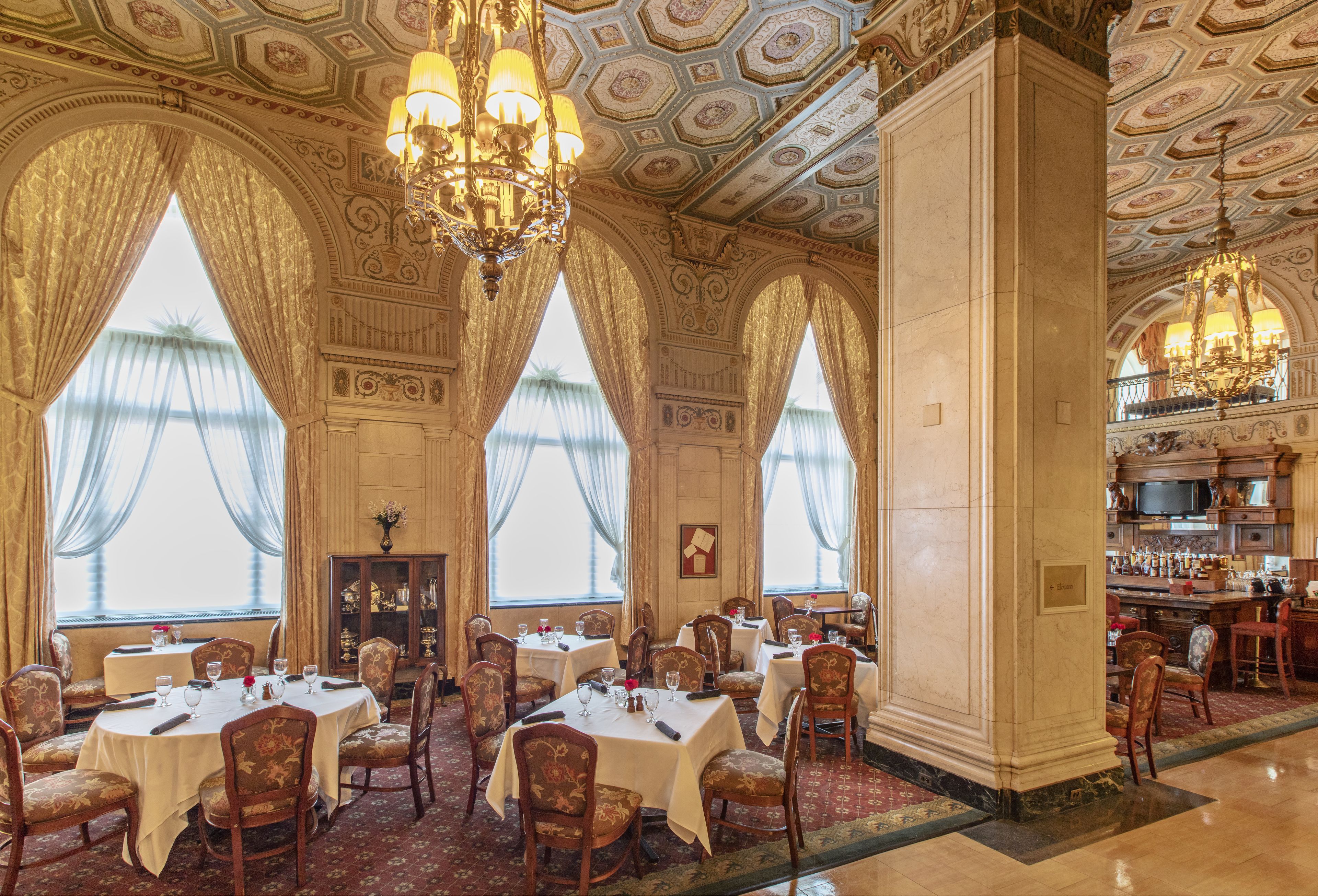 A grand dining room with ornate chandeliers, large windows with curtains, and a bar area.