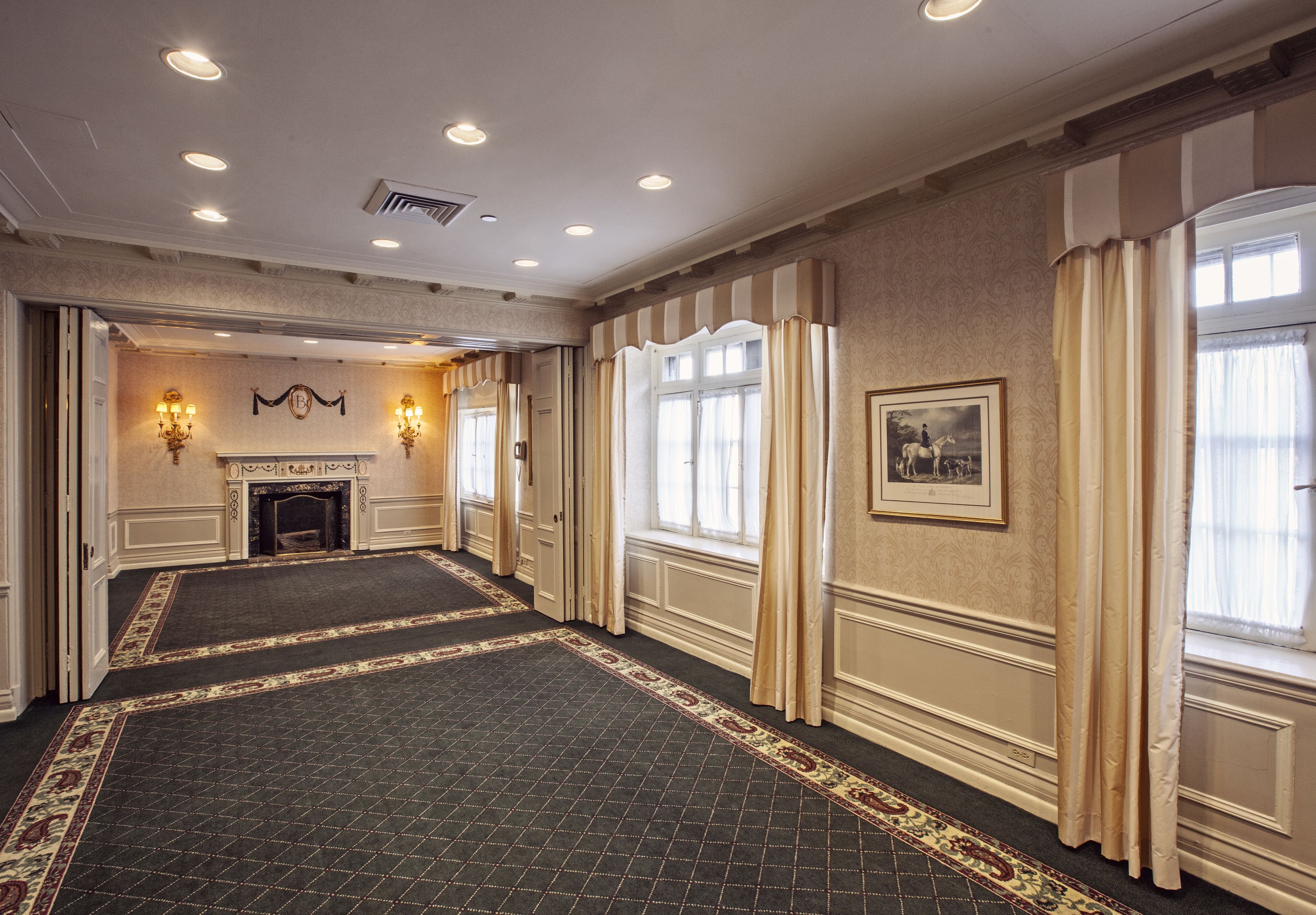 A spacious hotel lobby with a patterned carpet, a fireplace, and large windows with curtains.