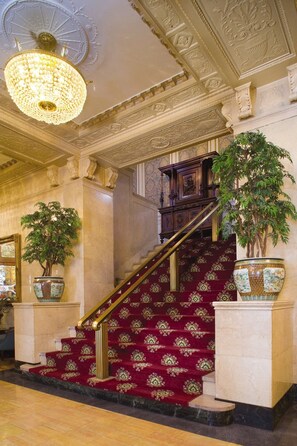 A grand staircase with red carpeted steps, flanked by large potted plants and a chandelier.