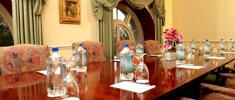 Un salón de conferencias con una larga mesa, sillas de estampado floral, botellas de agua y un jarrón con flores.