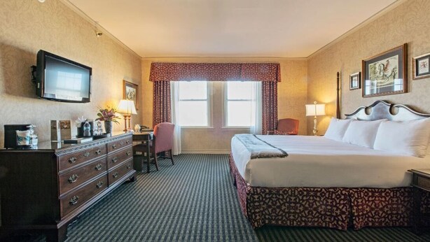 A hotel room with a large bed, a dresser, a desk, a television, and a window with curtains. - The Brown Hotel (Louisville)
