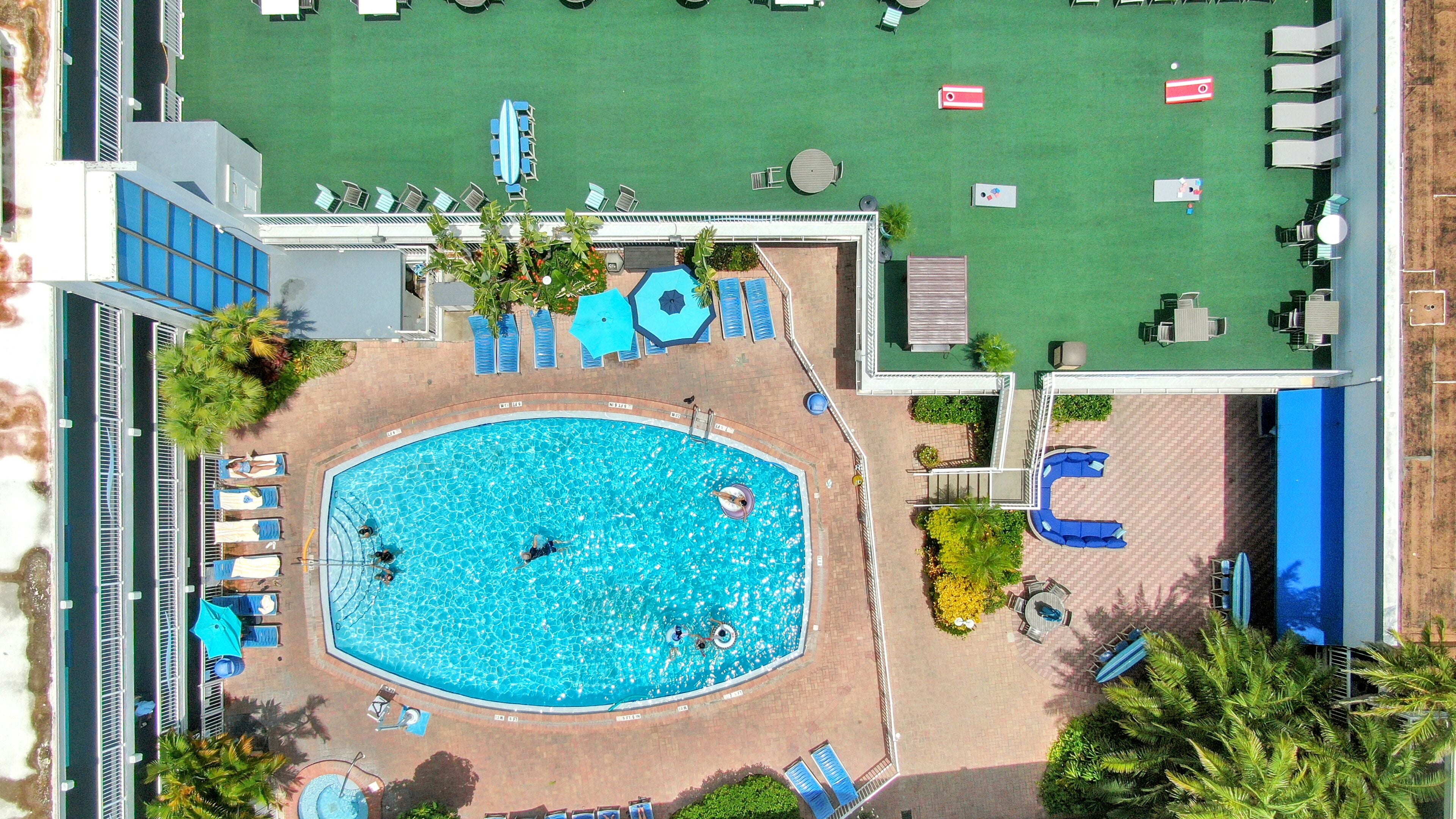 An aerial view of a hotel pool area with a central pool, surrounding lounge chairs, and a patio area with tables and chairs.