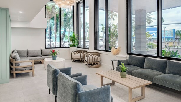 A modern hotel lobby with a grey sofa, wooden chairs, a round coffee table, and large windows offering a view of palm trees and buildings. - Bilmar Beach Resort (Treasure Island)