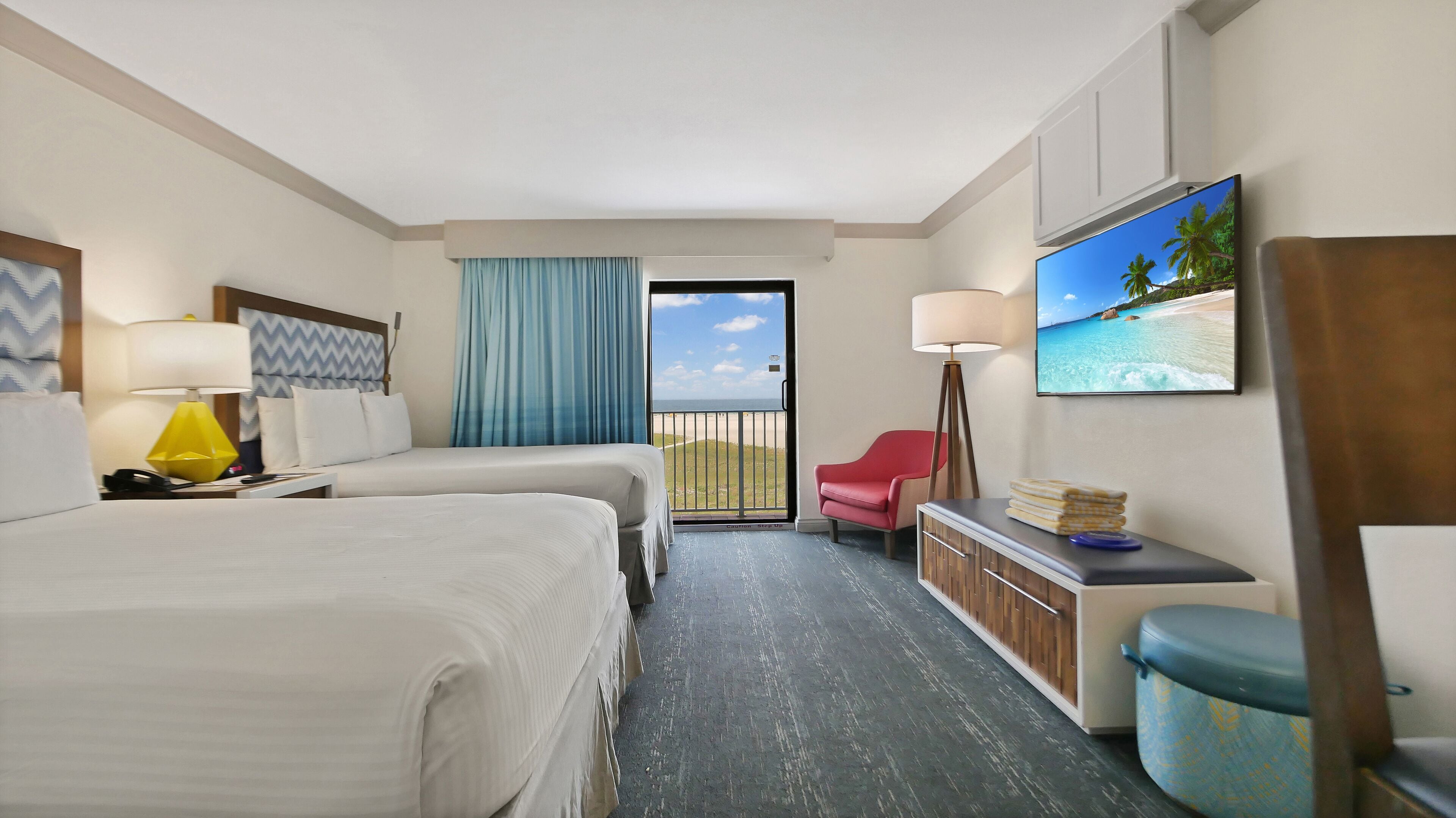 A hotel room with two beds, a desk, a chair, a TV, and a balcony with a view of the beach.