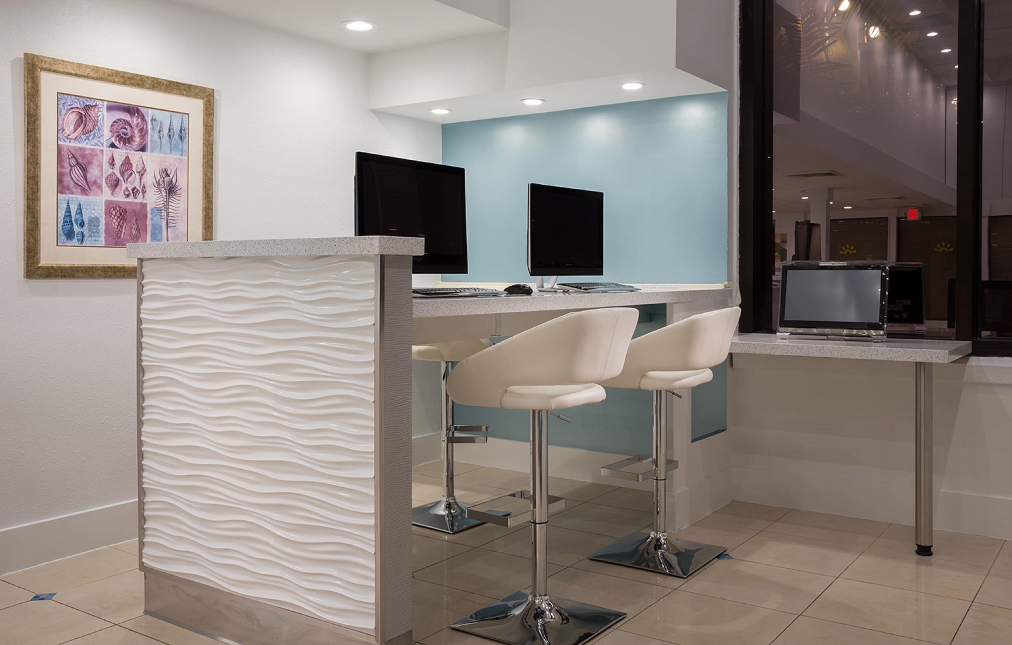 A modern reception area with a white textured partition, two bar stools, and a counter with two monitors.