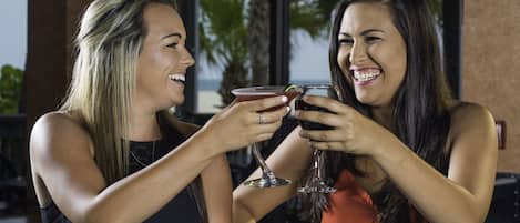Duas mulheres brindando com bebidas em uma mesa, em ambiente externo, com vista para palmeiras.