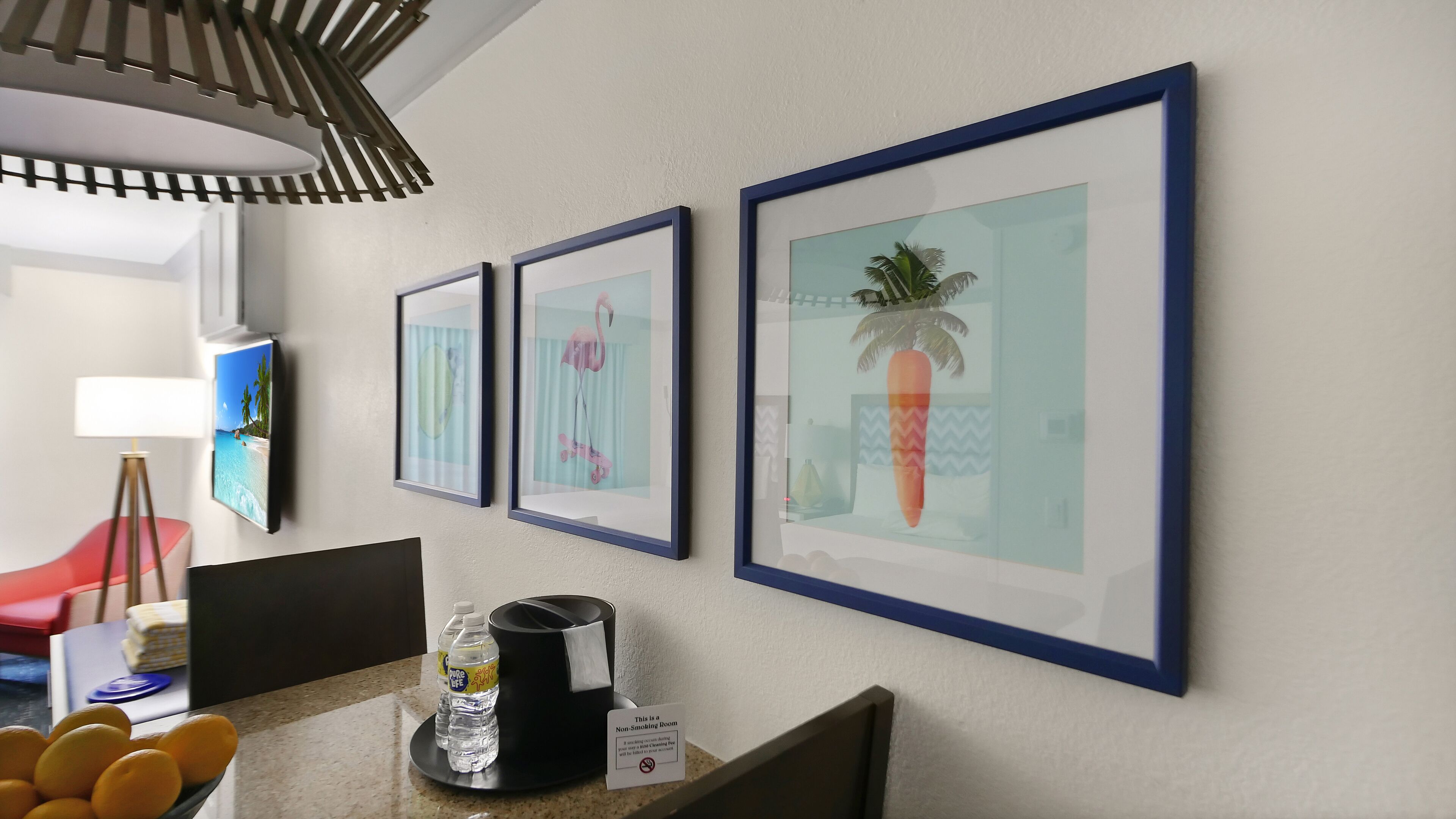 A hotel room with a dining area featuring a table with a coffee maker, water bottles, and a bowl of oranges. There are three framed pictures on the wall, and a television is mounted on the wall.