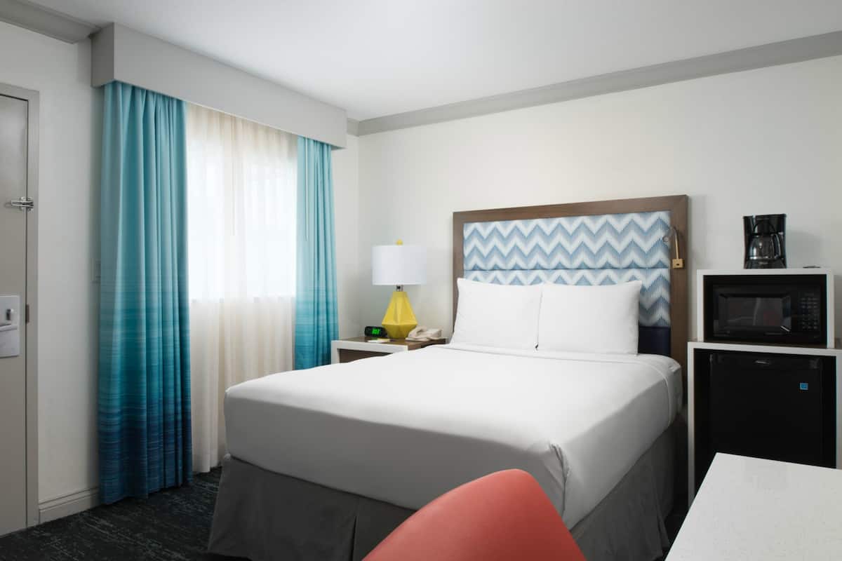 Quarto de hotel com cama, mesa de cabeceira com luminária, micro-ondas e cafeteira.