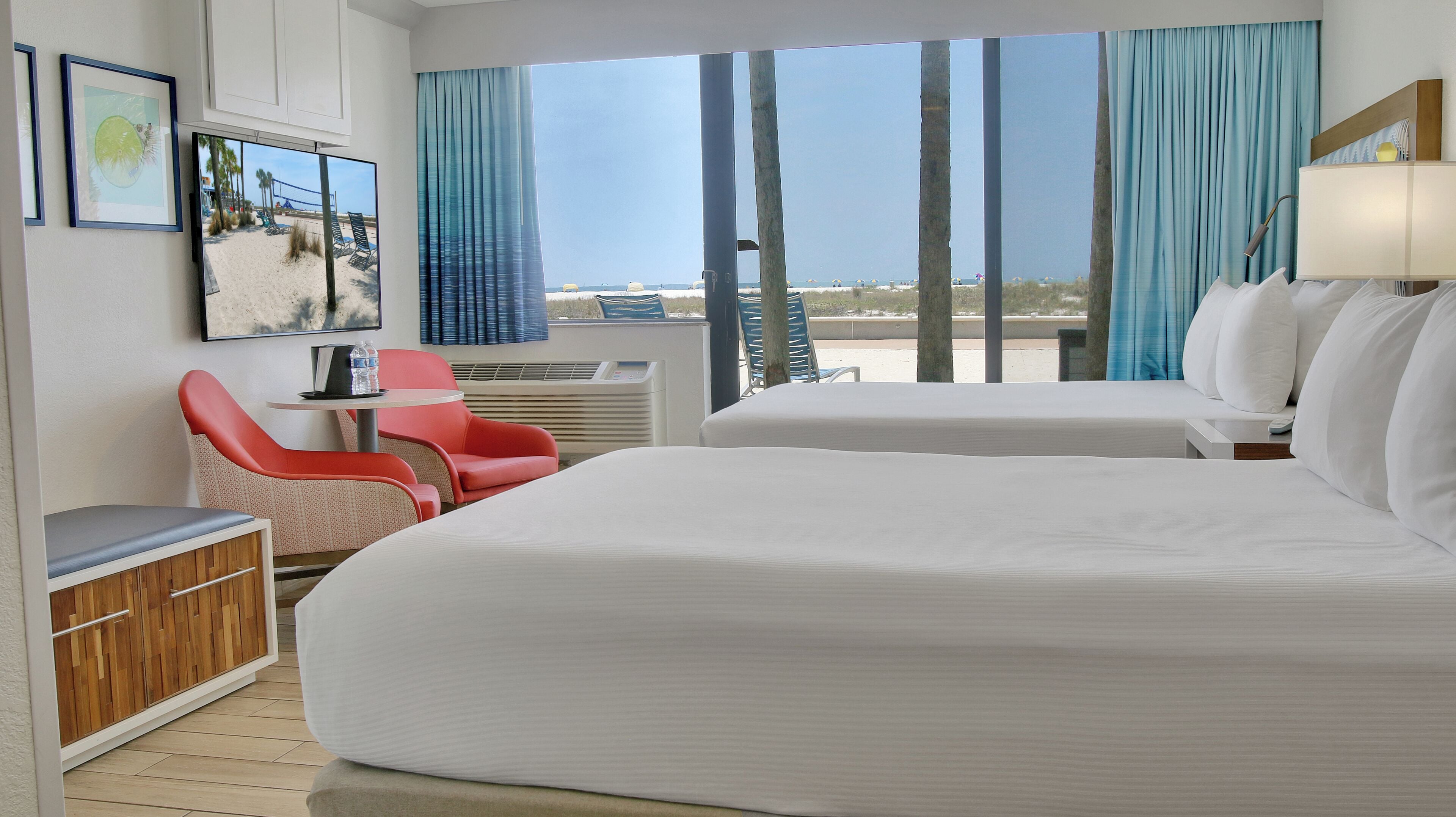 Habitación de hotel con dos camas, un escritorio, una silla y un amplio ventanal con vistas a la playa.