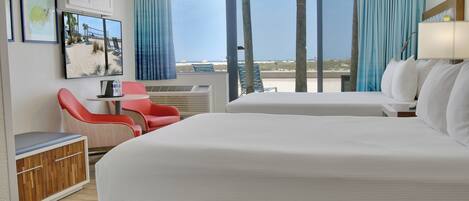 A hotel room with two beds, a desk, a chair, and a large window with a view of the beach.