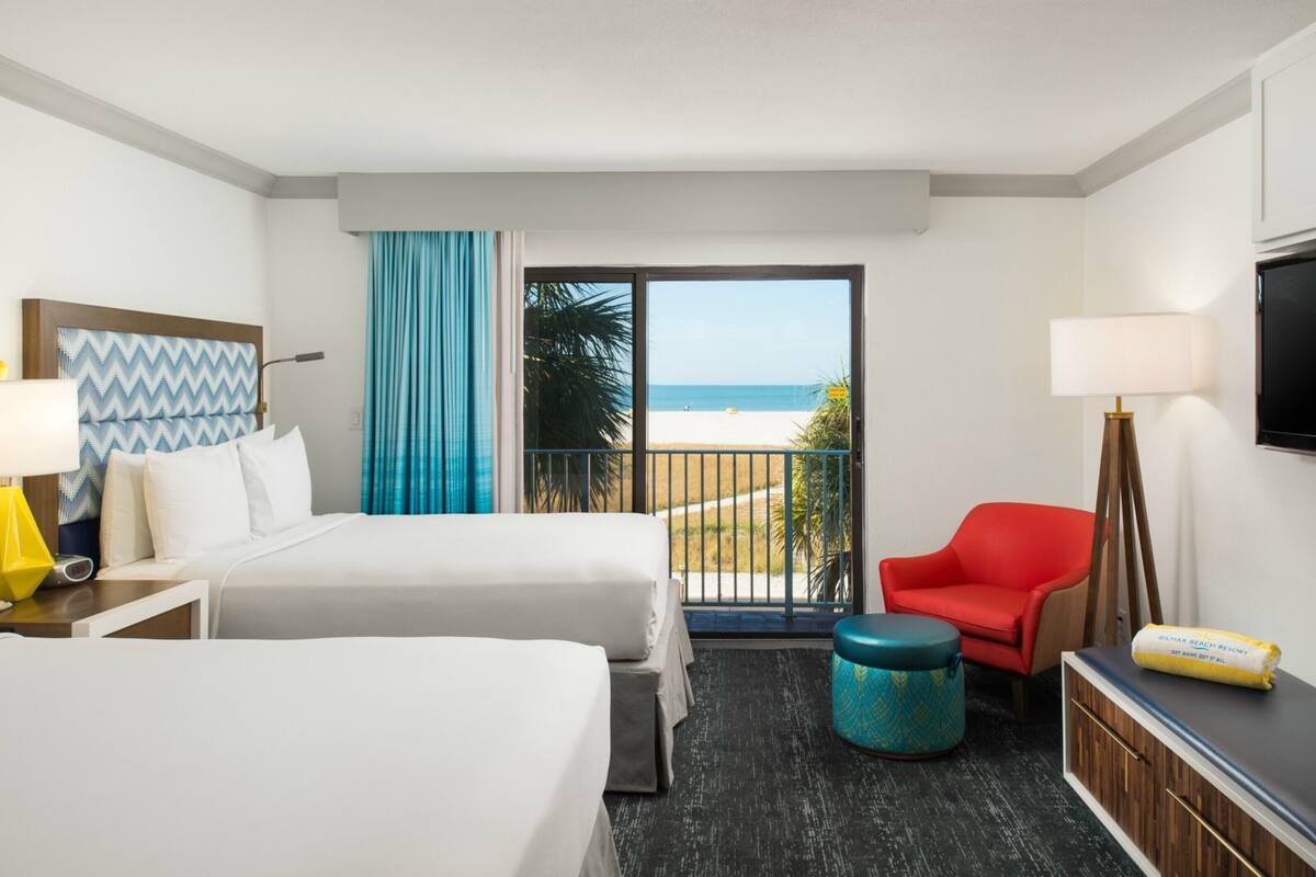 Quarto de hotel com duas camas, televisão, uma cadeira vermelha, uma mesinha e vista para a praia.